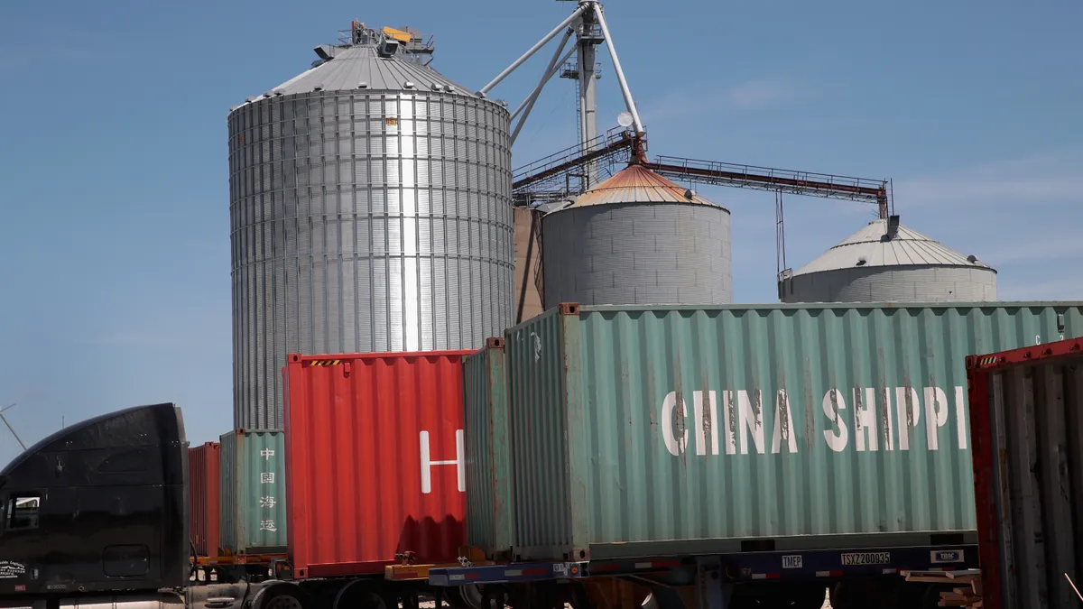 Shipping containers on a truck are loaded with soybeans as a grain elevator stands in the background